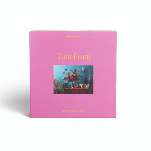 Display photo of the Tutti-Frutti puzzle. The box if a bright pink color with a photo of the completed puzzle on the front.  The puzzle features a photo of flowers and fruit on a table. 