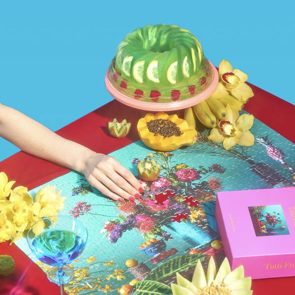 A person, with face off camera, works on the Tutti Fruitti puzzle.  The table, with puzzle almost complete, is full of fresh fruit and a lime jello mold cake. 