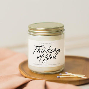 A glass jar candle with gold lid sits on a wooden candle coaster with wooden matches. The label reads "Thinking of You".
