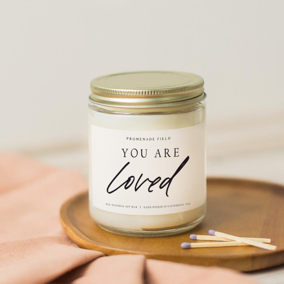 A glass jar candle with gold lid sits on a wooden candle coaster with wooden matches.  The label reads "You Are Loved". 