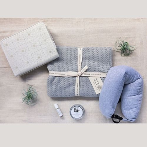A display photo of the Extended Hospital Stay Gift Box picturing a throw, next wrap, body balm, aromatherapy rollerball, grey throw, and bedside pocket.