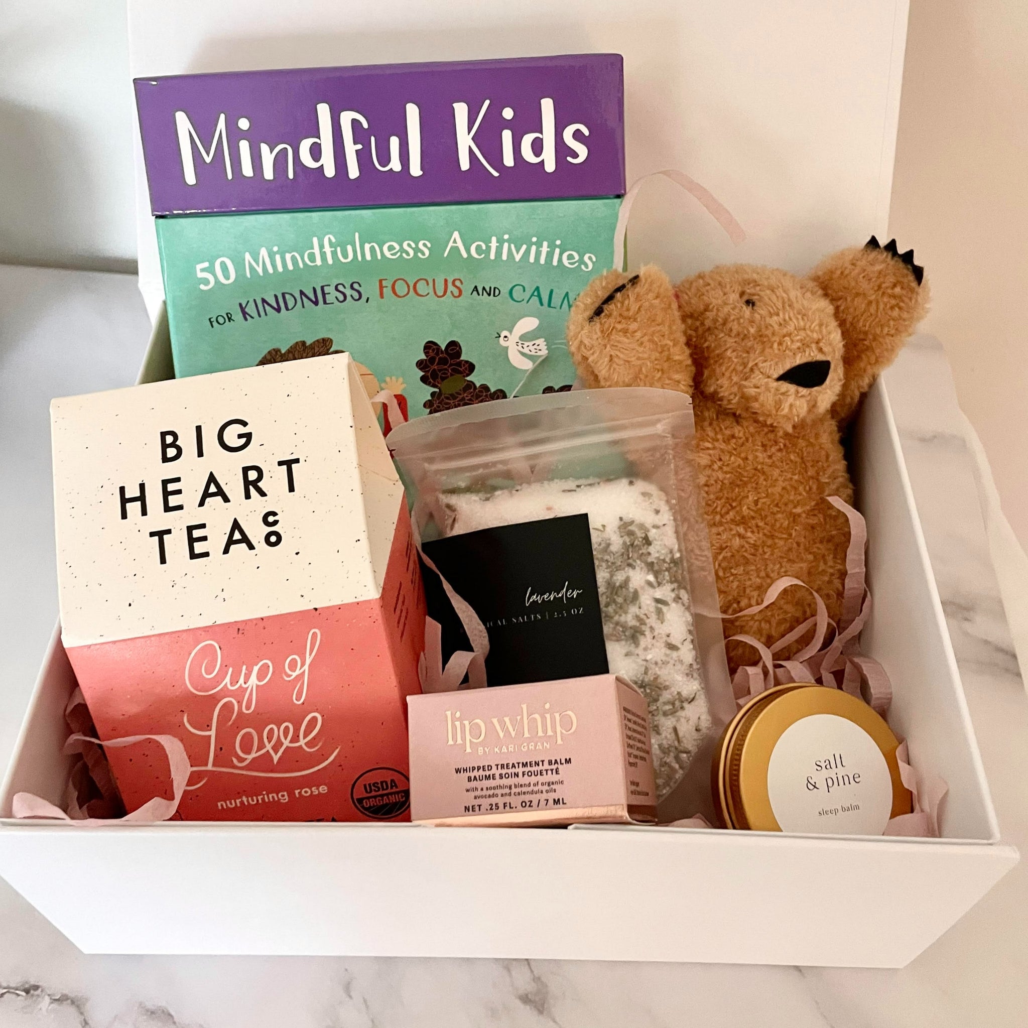 picture featuring a family support themed care package for families going through difficult chapters of life. Included in the gift are mindfulness cards for kids, a small plush bear, cup of love tea, lip whip moisturizing lip balm, lavender bath salts and sleep balm